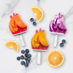Three unicorn-shaped popsicles—red, orange, and purple—sit on a marble surface with blueberries and orange slices, made using Stackable Pop Molds for easy homemade treats.