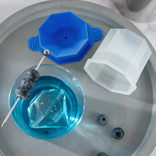 A blue drink with a large diamond-shaped ice cube made using tovolo Diamond Ice Molds (Set of 2) is garnished with blueberries, shown beside blue and clear molds and loose blueberries on a gray tray.