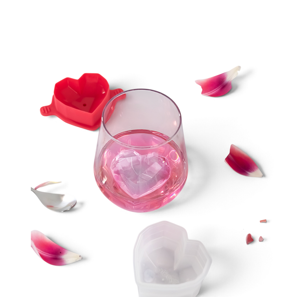 A glass of pink drink with a large heart-shaped ice cube sits on marble next to pink petals, a spoon, and tovolo Heart Ice Molds (Set of 2), perfect for creating charming heart-shaped cubes.