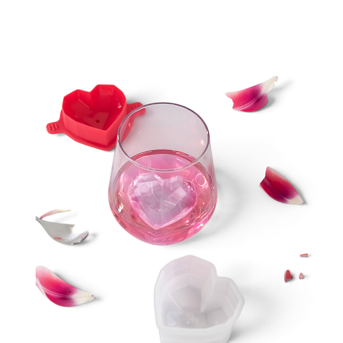 A glass of pink drink with a large heart-shaped ice cube sits on marble next to pink petals, a spoon, and tovolo Heart Ice Molds (Set of 2), perfect for creating charming heart-shaped cubes.