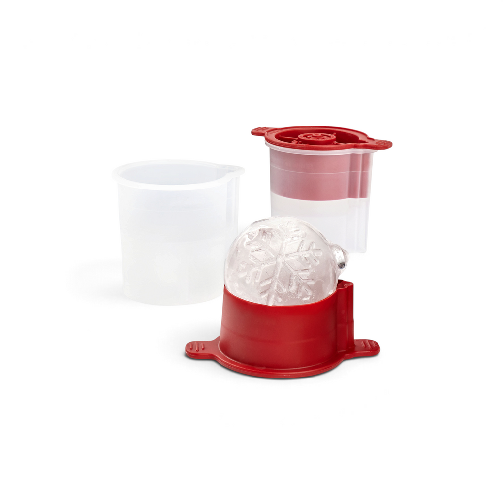 The tovolo Holiday Ice Molds, featuring red and clear plastic parts, sit beside a snowflake-patterned ice sphere—perfect for festive cocktails and a great Christmas gift idea. Another clear cup appears in the background.