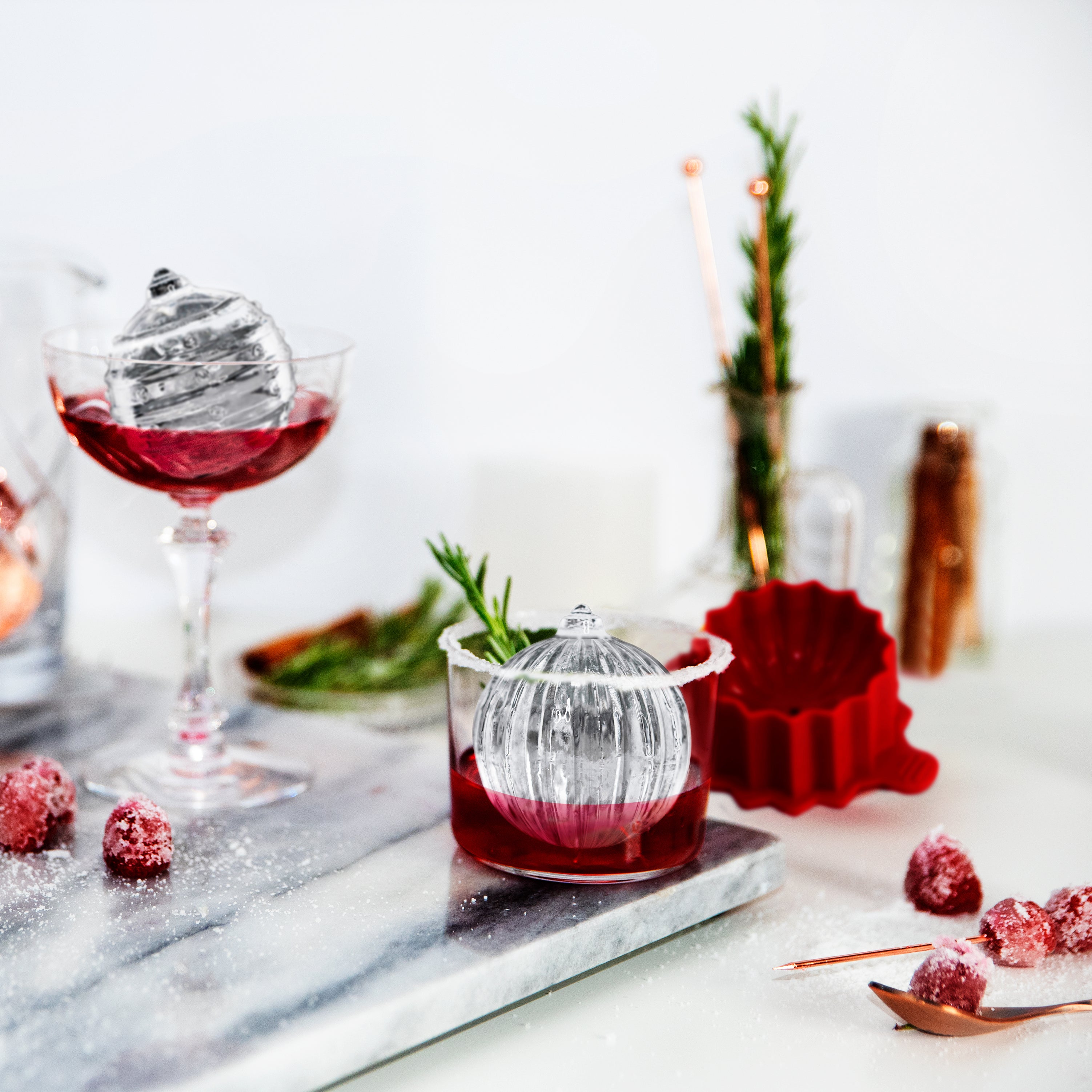 A festive display features cocktails with ornament-shaped spheres made using tovolo's Dots and Stripes Ornament Ice Molds (Set of 2), garnished with herbs and cranberries—perfect for a stylish Christmas gift.