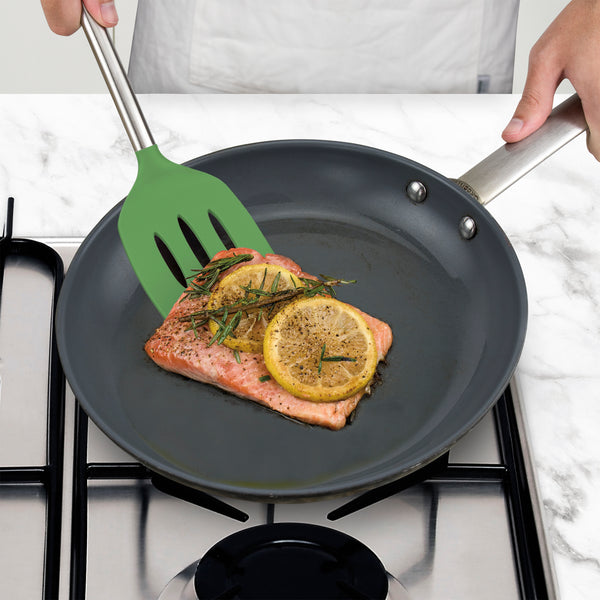 A person uses a Silicone Slotted Turner to cook a piece of salmon with lemon slices and herbs in a frying pan on the stovetop.