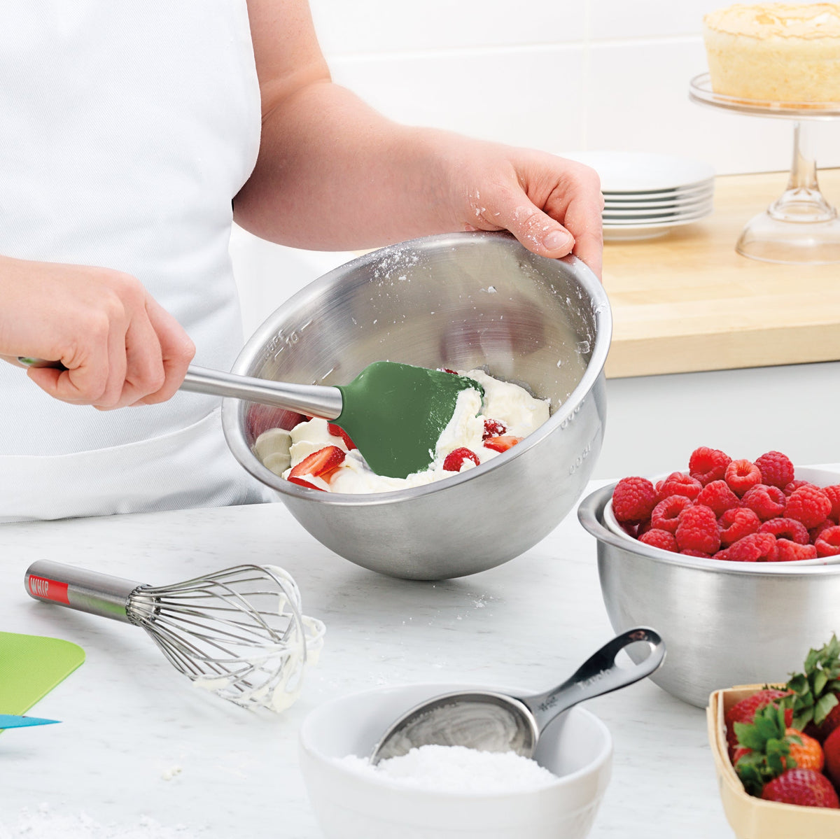 A person mixes whipped cream and strawberries in a metal bowl using a green Flex-Core Spatula. Nearby are a bowl of raspberries, a whisk, a sifter with powdered sugar, and a cake on a stand in the background.