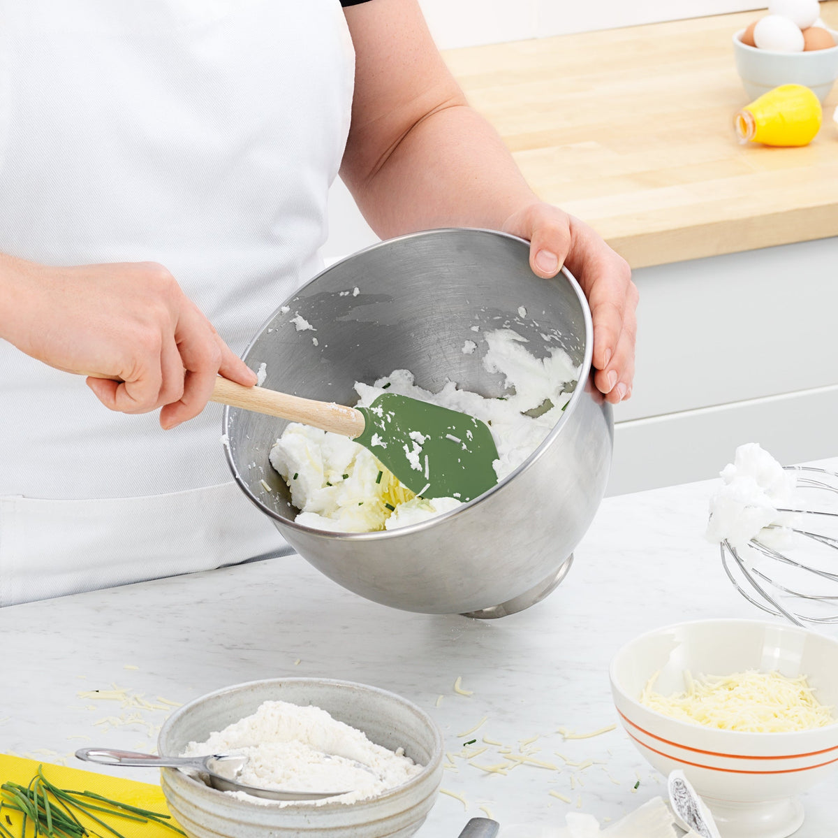 Wearing a white apron, someone uses a green Flex-Core Spatula to mix ingredients in a metal bowl, with flour, grated cheese, and chives visible on the counter.