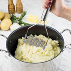 A hand mashes potatoes in a pot using a Silicone Potato Masher. Nearby on a marble countertop are whole potatoes, butter, garlic, and herbs.