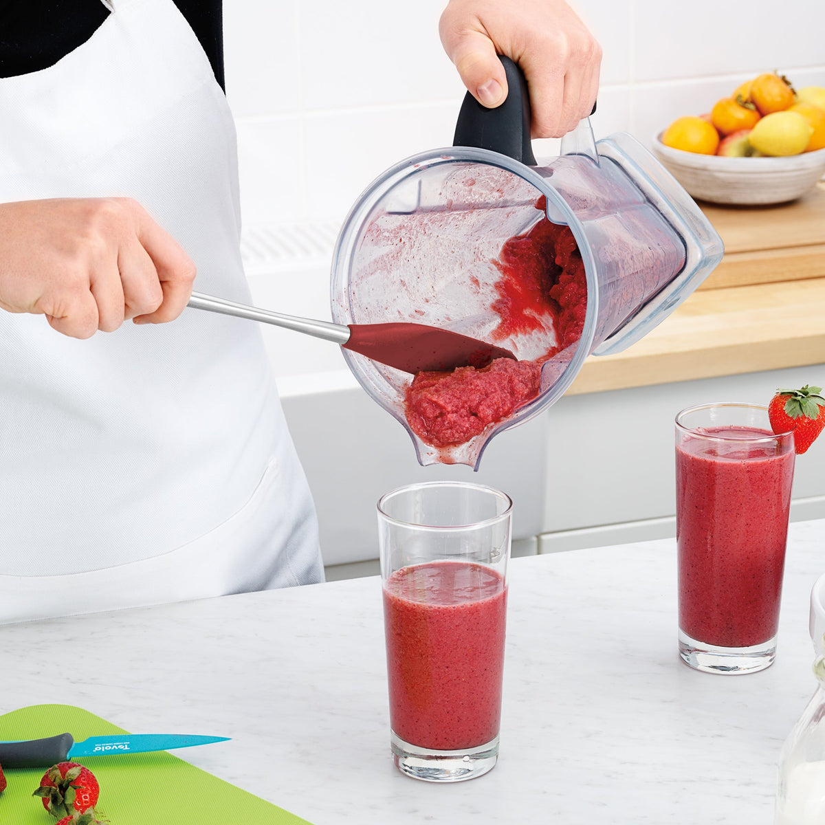 Wearing a white apron, someone uses the tovolo Flex-Core Silicone Jar Scraper to pour a thick red smoothie from a blender into a tall glass on the kitchen counter. Another filled glass, strawberries, a knife, and fruit bowl are also present.