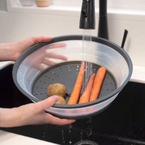A person uses the Collapsible Microwave Food Cover as a strainer under running water in a sink, rinsing two potatoes and three carrots, while water drains through its bottom holes.