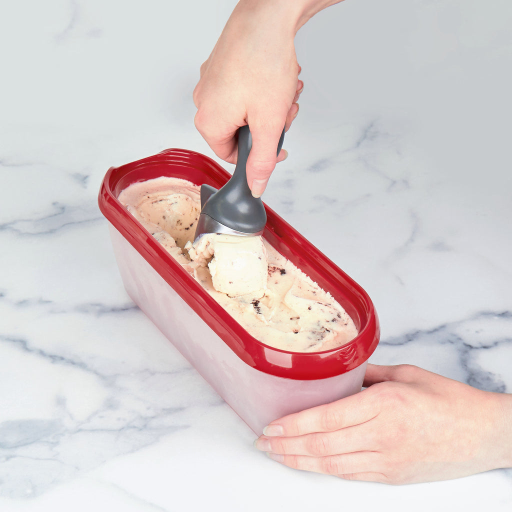 A person uses a gray ice cream scoop to serve ice cream from a red Glide-A-Scoop Ice Cream Tub on a marble countertop.