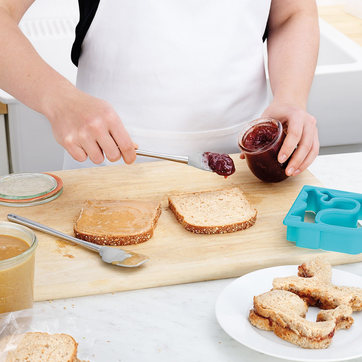 Using the tovolo Flex-Core Silicone Mini Spatula + Spoonula, someone spreads jelly on bread next to a peanut butter slice; jars, knives, a blue sandwich cutter, and shaped sandwiches sit on a wooden board and counter.