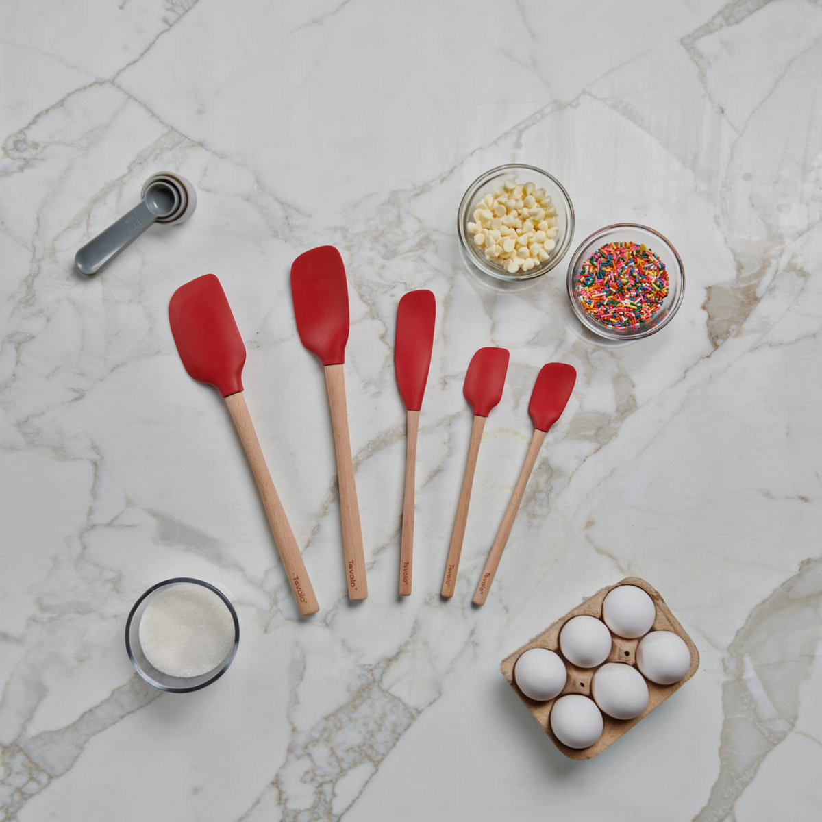 Five Flex-Core Wood-Handle Spatulas in red, each a different size, are arranged on a marble countertop with eggs, white sugar, white chocolate chips, sprinkles, and a metal tablespoon.