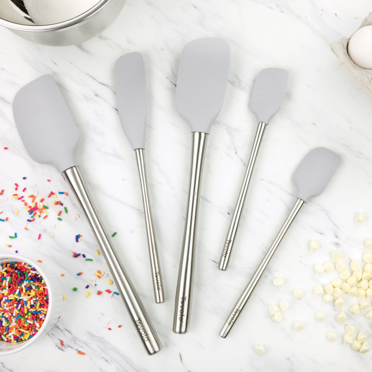 Five Flex-Core All-Silicone Spatula Set of 5 pieces in gray are arranged on a marble countertop alongside sprinkles, white chocolate chips, eggs, and a metal mixing bowl.