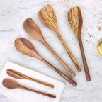 Five Olivewood Utensils, including a spatula and pancake turner, are arranged on a marble surface beside a white cloth, scattered grains, and a glass of oil. These nonstick-safe utensils bring elegance to any kitchen.