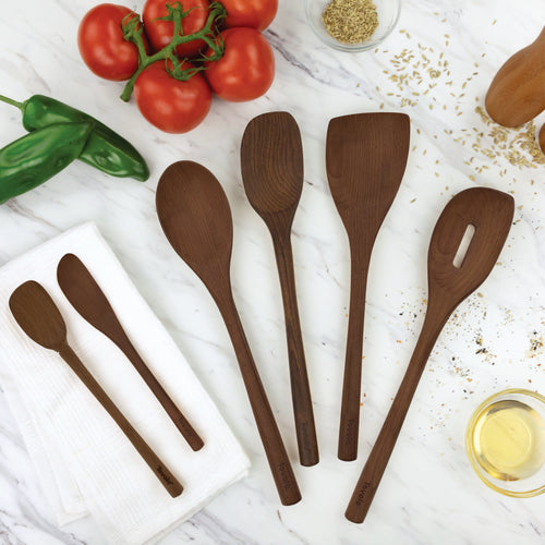 Five Beechwood Utensils, including an angled turner, are arranged on a marble countertop with fresh tomatoes, green peppers, a white towel, a small bowl of oil, and grains—ideal for non-stick cookware.