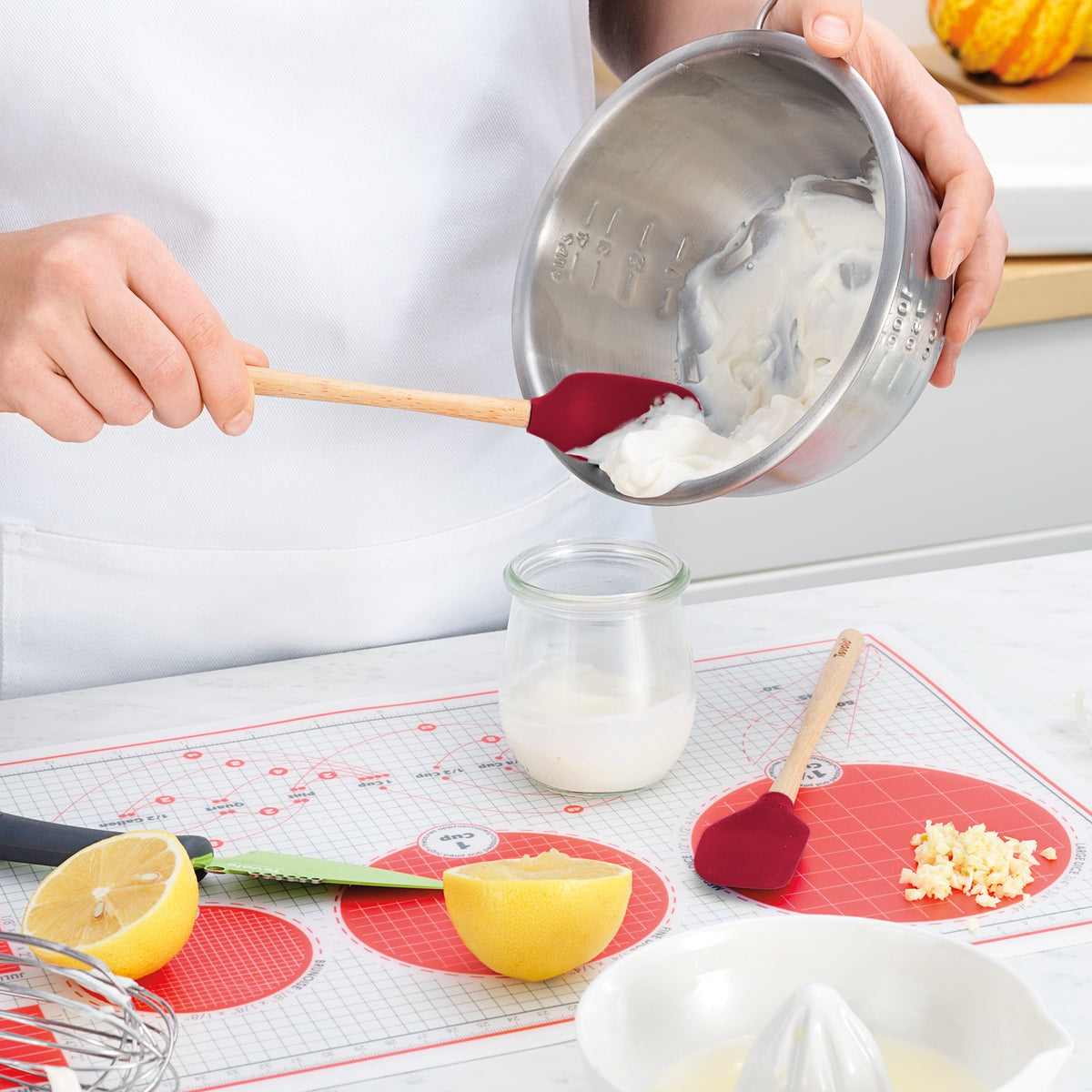 Someone uses a tovolo Flex-Core Silicone Mini Spatula + Spoonula in red to transfer a creamy mixture from a metal bowl into a glass jar. Nearby are a halved lemon, knife, minced garlic, and another red spatula on a red and white mat.