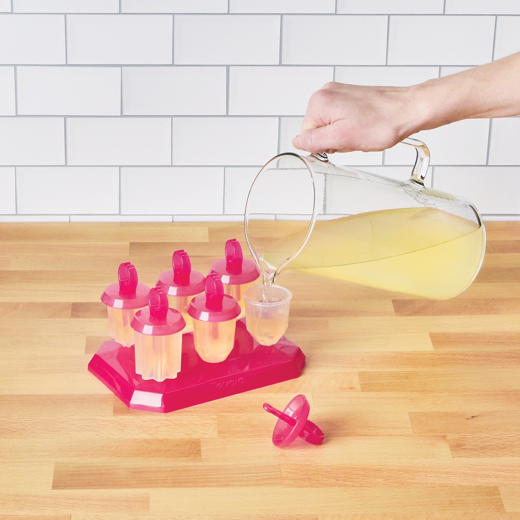 A hand pours yellow liquid from a glass pitcher into Jewel Pop Molds on a slim tray atop a wooden counter, with several pink-lidded molds already filled, one lid beside the tray, and a white tiled wall in the background.