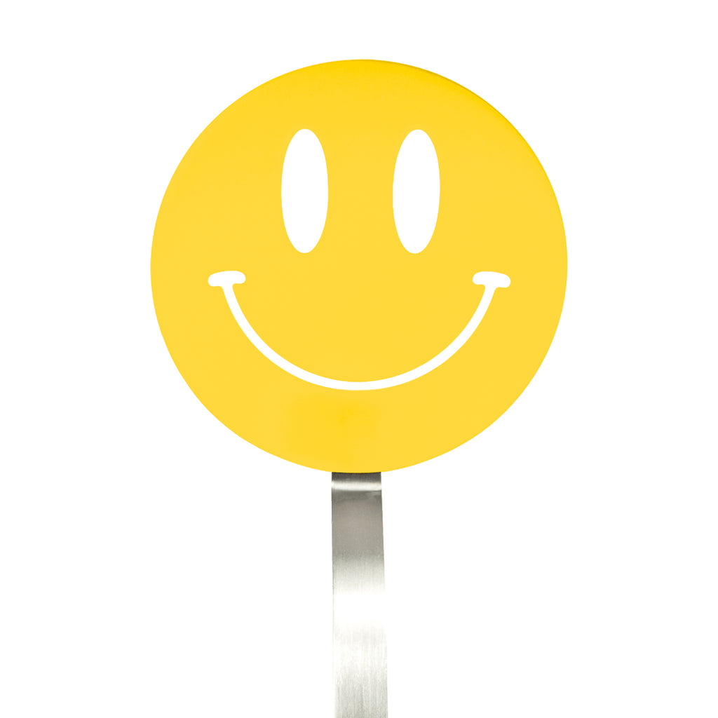 A yellow smiley face sign with a wide, curved smile and oval eyes is mounted on a silver pole, resembling the cheerful design of the Spatulart Flex Turner kitchen tool, which is safe for use with nonstick skillets.