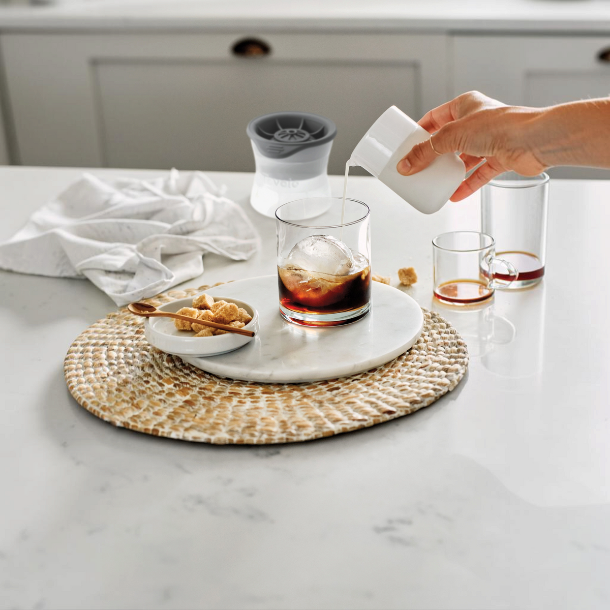 A hand pours milk from a small pitcher into iced coffee with slow-melting ice made using tovolo Sphere Ice Molds (Set of 2); on the white table are brown sugar cubes, a spoon, and syrup.