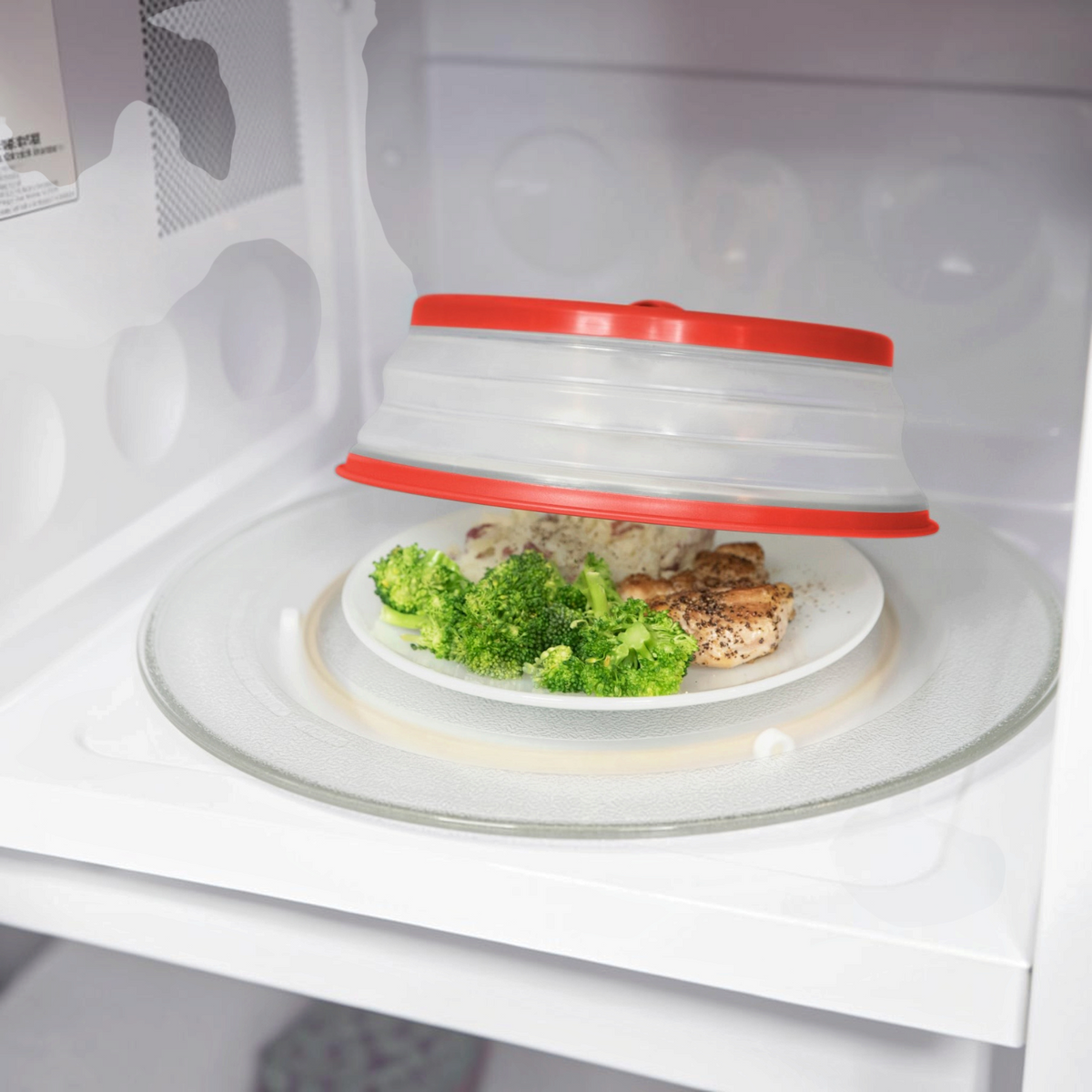 A plate of cooked chicken, broccoli, and rice sits in the microwave, covered by a partially lifted Collapsible Microwave Food Cover.