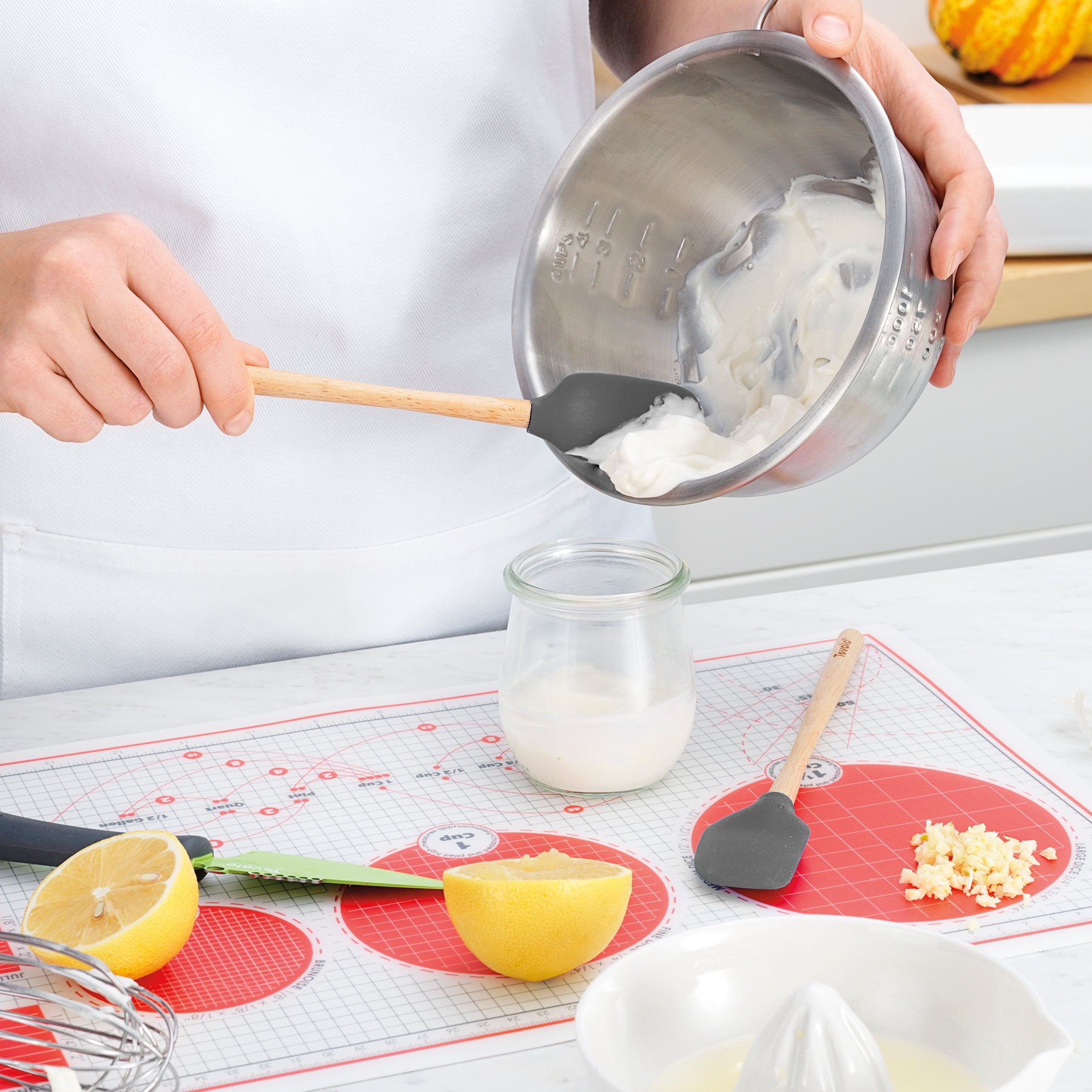 Using the tovolo Flex-Core Silicone Mini Spatula + Spoonula, a person transfers a creamy mixture from a metal bowl into a glass jar on a kitchen counter with a halved lemon, knife, whisk, minced garlic, and red silicone mat.