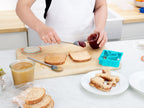 A person uses a tovolo Flex-Core Mini Spatula + Spoonula with Stainless Steel Handle to spread jelly onto peanut butter bread on a cutting board, with sandwich pieces, cookie cutters, and jars of peanut butter and jelly nearby.