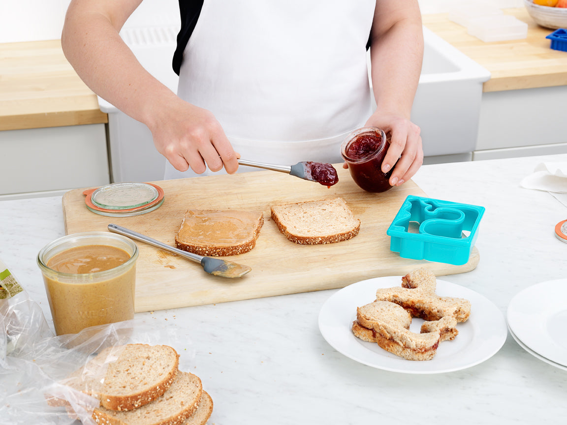 A person uses a tovolo Flex-Core Mini Spatula + Spoonula with Stainless Steel Handle to spread jelly onto peanut butter bread on a cutting board, with sandwich pieces, cookie cutters, and jars of peanut butter and jelly nearby.