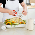 Wearing a white apron, someone uses Kitchen Shears to cut twine while prepping a raw, herb-seasoned chicken in a roasting pan with squash and herbs. A spool of twine and a jar of salt rest on the table nearby.