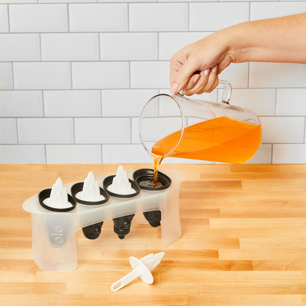 A hand pours orange liquid from a glass pitcher into Penguin Pop Molds on a wooden countertop, with a white tiled wall in the background—perfect for making homemade frozen treats.