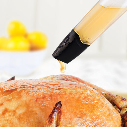 A close-up of a turkey being basted with the Bulb Baster, featuring a silicone bulb and dripless design, as golden juices coat the browned, roasted skin; blurred lemons appear in the background.