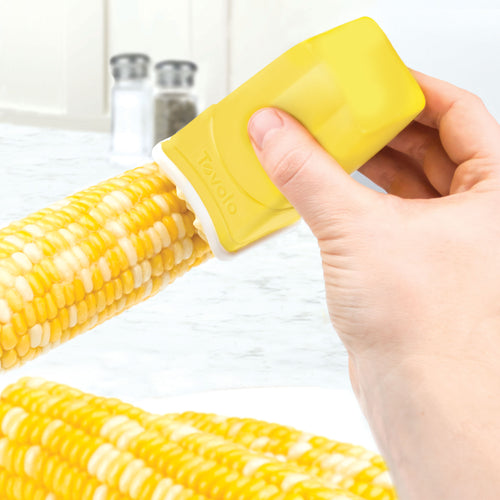 A hand uses the Butter Sleeve to spread butter evenly on corn; salt and pepper shakers are blurred in the background on a kitchen counter.