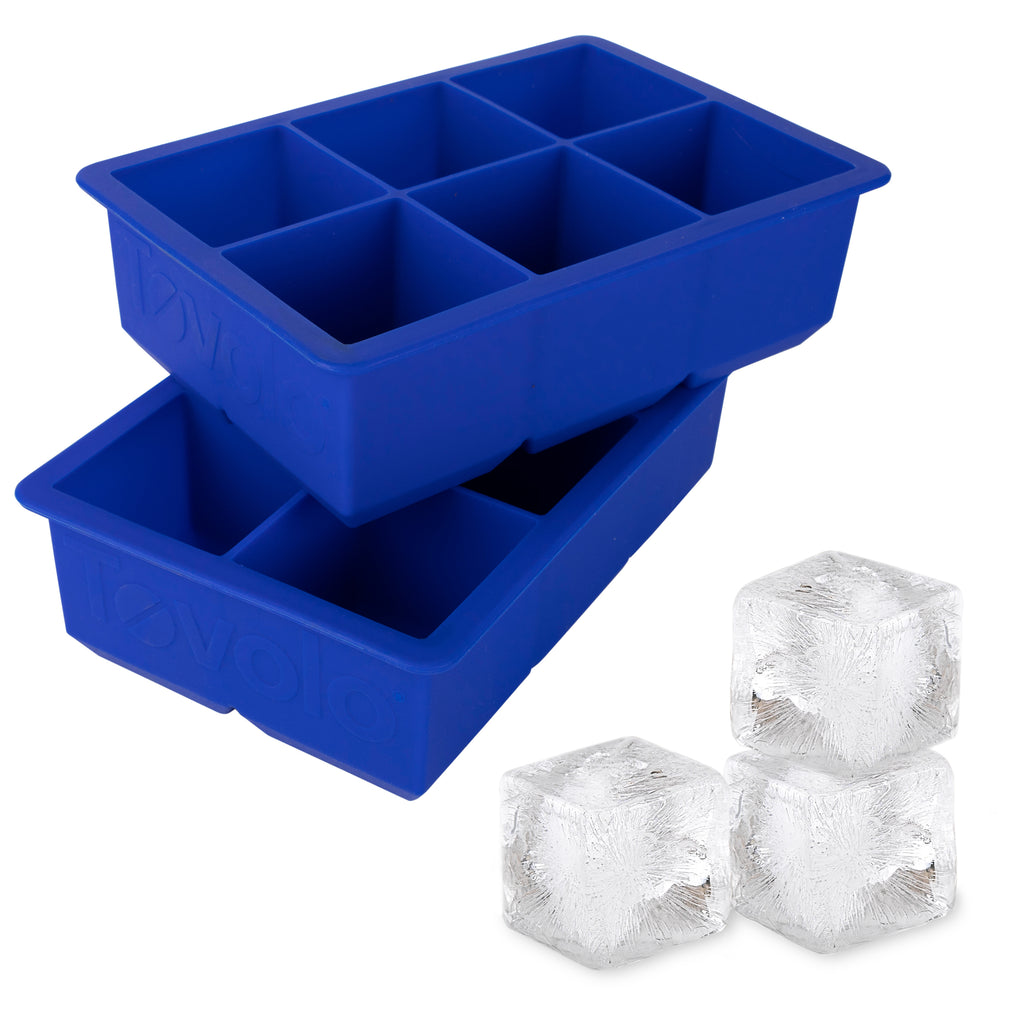 The tovolo King Cube Silicone Ice Molds (Set of 2) are stacked, each featuring six large compartments, with three clear craft ice cubes in front—ideal for chilling drinks without dilution.