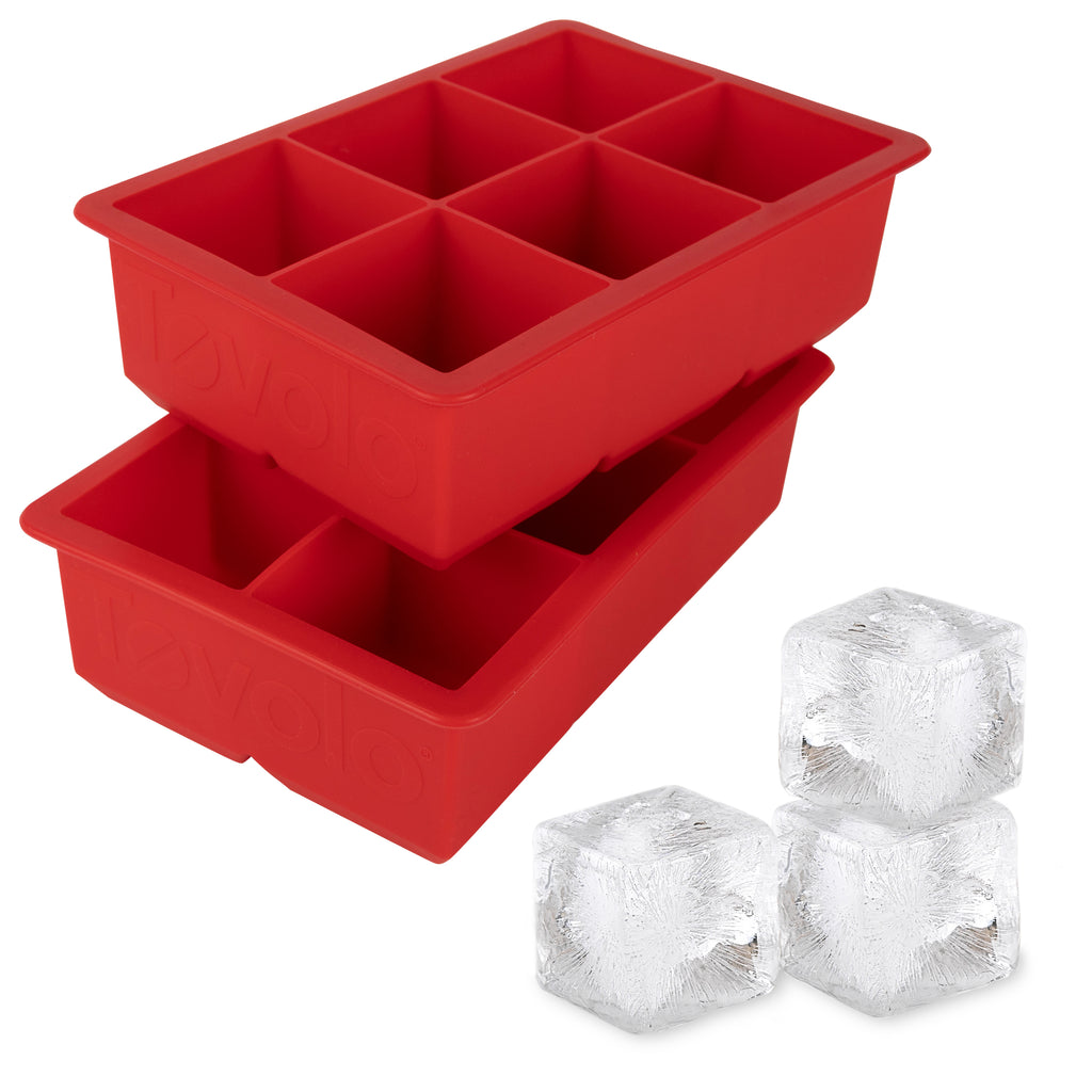 Two red tovolo King Cube Silicone Ice Molds (Set of 2), each with six large square compartments, are stacked. In front, three large clear craft ice cubes are displayed.