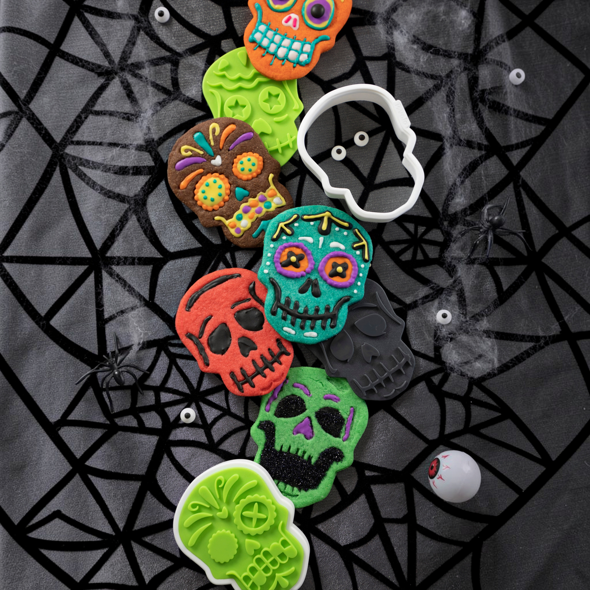 Colorful skull cookies made with Skull Cookie Cutters are decorated with icing and sprinkles on a spiderweb-patterned surface, surrounded by fake eyeballs, spiders, and a white skull-shaped cookie cutter.