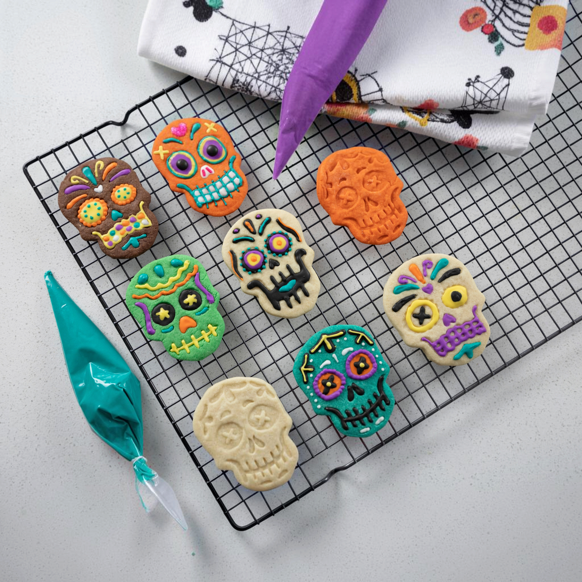 Sugar skull-shaped cookies, made with Skull Cookie Cutters, rest on a cooling rack, their bright icing reminiscent of festive Christmas cookies. A nearby piping bag of blue icing is ready to add more creative designs.