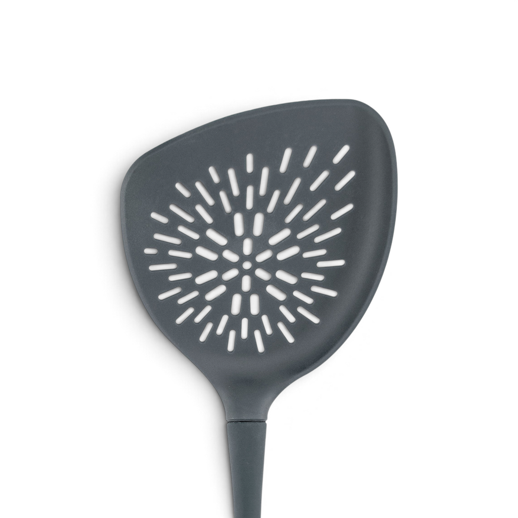 The Roasted Veggie Tool is a black non-stick spatula with a wide triangular head and sunburst-pattern holes for easy draining—ideal as a veggie turner when serving roasted vegetables.