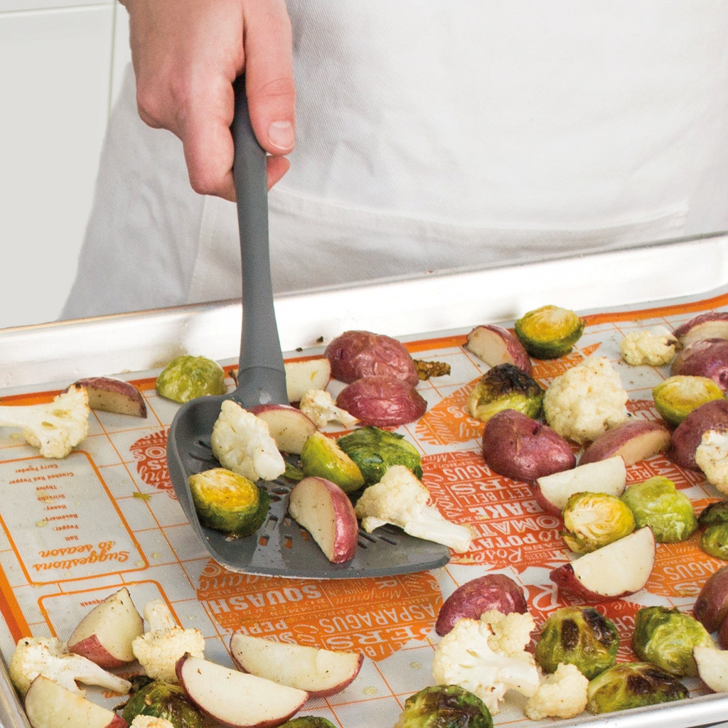 Wearing a white apron, a person uses the Roasted Veggie Tool to scoop Brussels sprouts, red potatoes, and cauliflower from a baking sheet lined with an orange silicone mat.