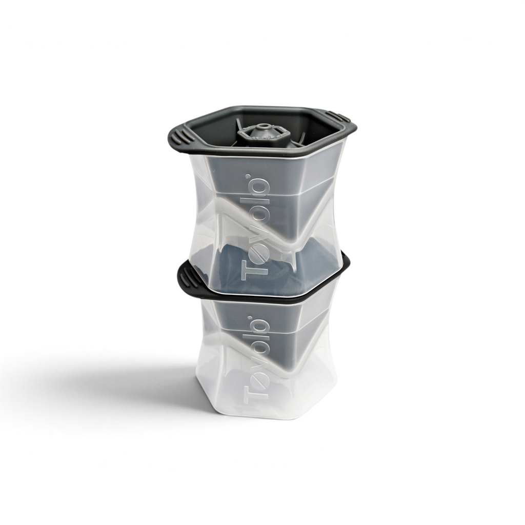 Two stacked tovolo Colossal Ice Molds (Set of 2) in clear and dark gray create large, geometric, slow-melting ice cubes. Each mold features the tovolo logo on the side.