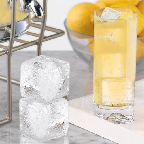 A glass of sparkling drink with ice cubes sits on a tray beside two stacked Colossal Ice Molds by tovolo. In the background, a bowl of lemons and a beverage dispenser highlight the Colossal Ice Molds (Set of 2) in use.