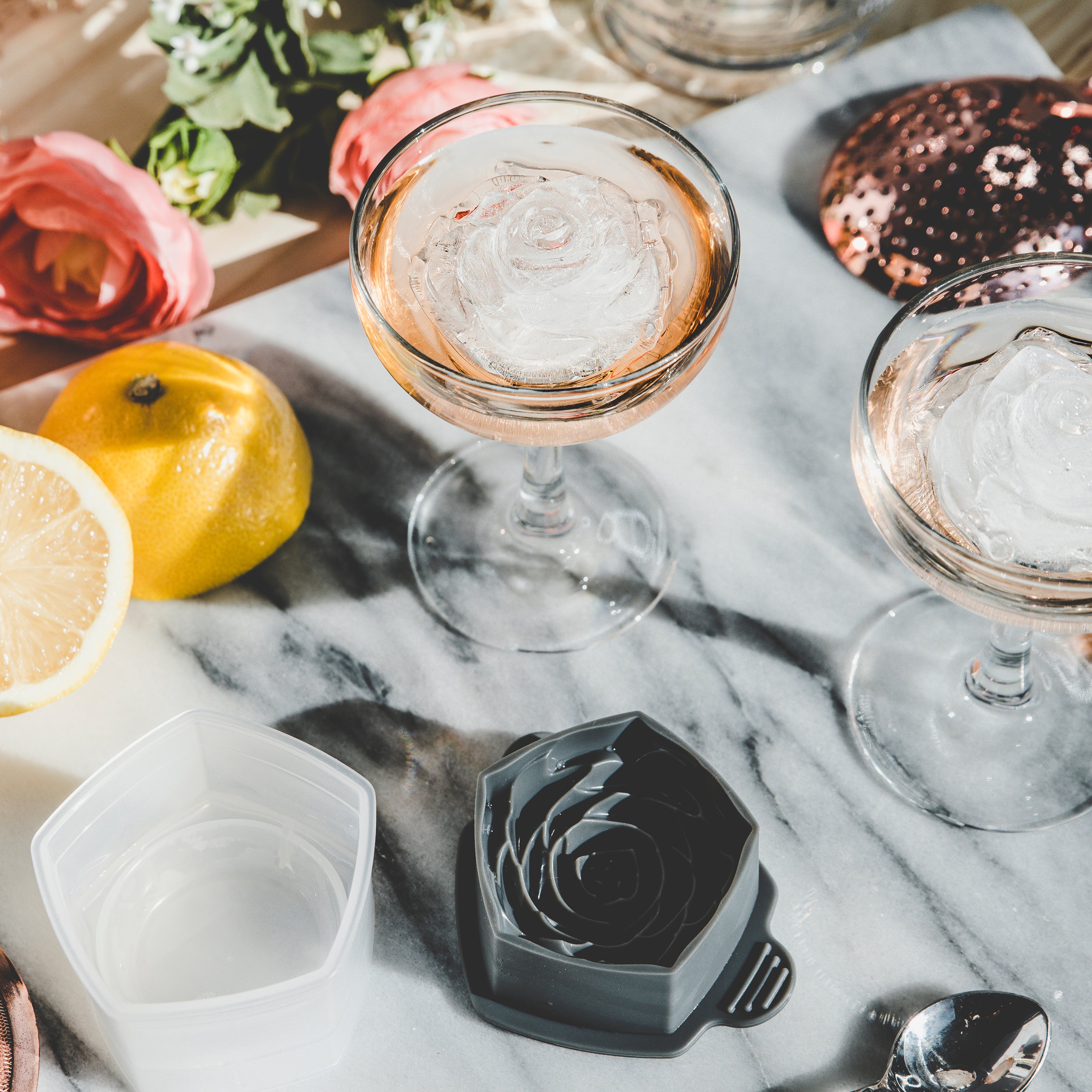 Two coupe glasses with drinks and rose-shaped ice from the tovolo Rose Ice Mold (Set of 2) rest on a marble surface, surrounded by lemon slices and flowers, while sunlight casts soft shadows over the scene.