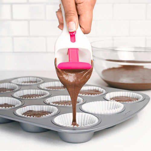 A hand uses the Cupcake Scoop to pour chocolate cake batter into paper-lined muffin cups, while a glass bowl with extra batter sits on the kitchen counter in the background.