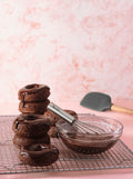 A stack of chocolate-glazed donuts sits on a cooling rack next to a glass bowl of chocolate glaze, with the Mini Whisk ready for quick whisking. A gray spatula rests in the background on a pink surface with a marbled pink and white backdrop.