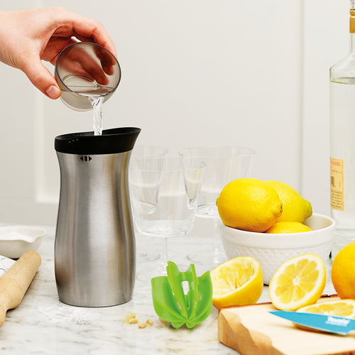 A hand pours liquid from a metal jigger into a stainless steel Cocktail Shaker on a kitchen counter with lemons, a citrus squeezer, cutting board, knife, glasses, and a bottle—ideal for mixing professional-quality cocktails.