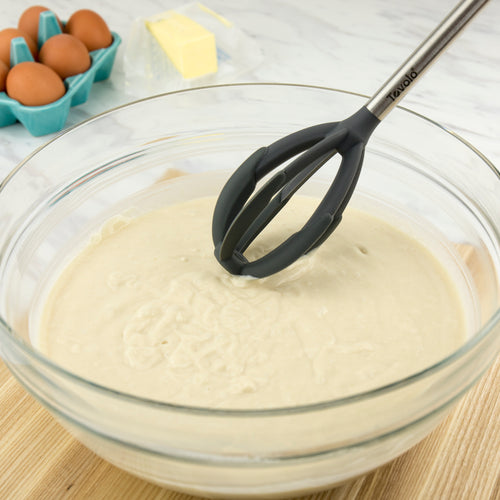 A glass bowl with cake batter is being mixed using the Better Batter Tool. In the background, a carton of brown eggs and a stick of butter sit on the countertop.